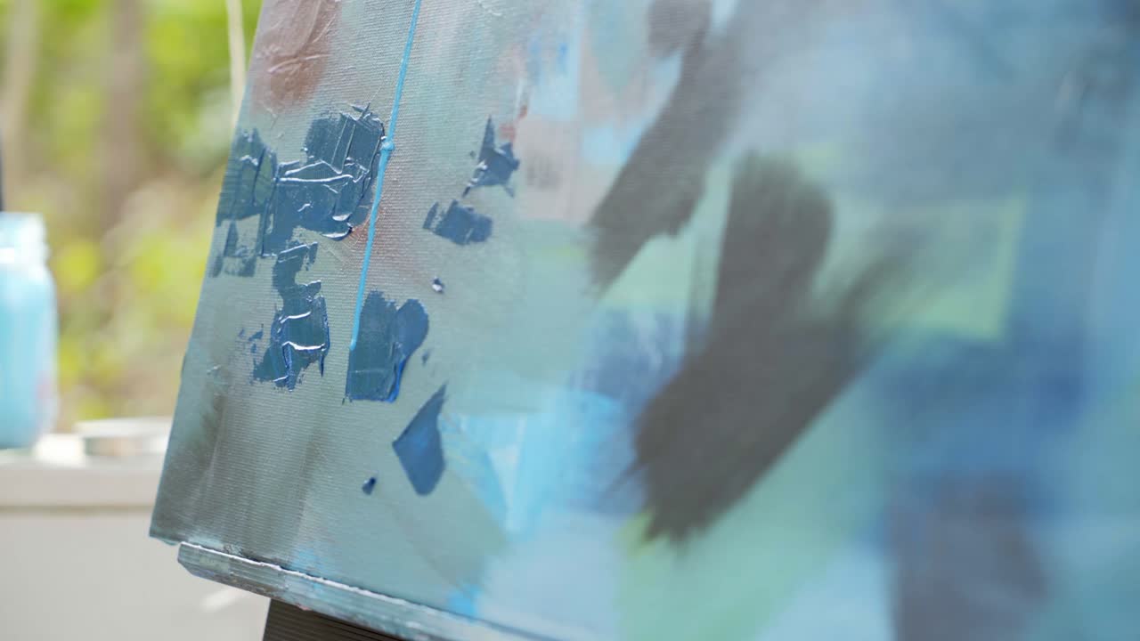 Blue paint dripping down canvas of abstract multicolored painting outside during the day, camera tilt down