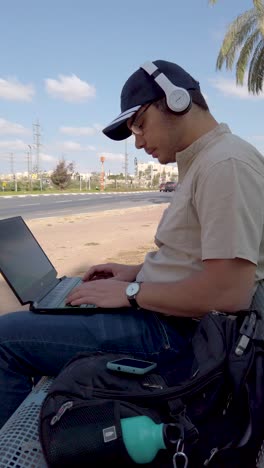 A man is sitting on a bench typing on a laptop at park, remote work.