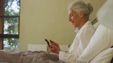 Ältere-Frau-Mit-Gemischter-Abstammung-Liegt-Mit-Smartphone-Im-Bett-Und-Lächelt