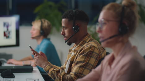 Man-In-Business-Team-Wearing-Headsets-Looking-At-Mobile-Phone-Working-Late-In-Sales-Or-Support