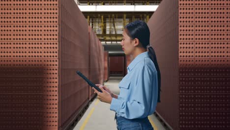 side view of asian female professional worker with red brick packed in stacks are stored, observes by looking up before she come to concentrating on the tablet and keep on checking