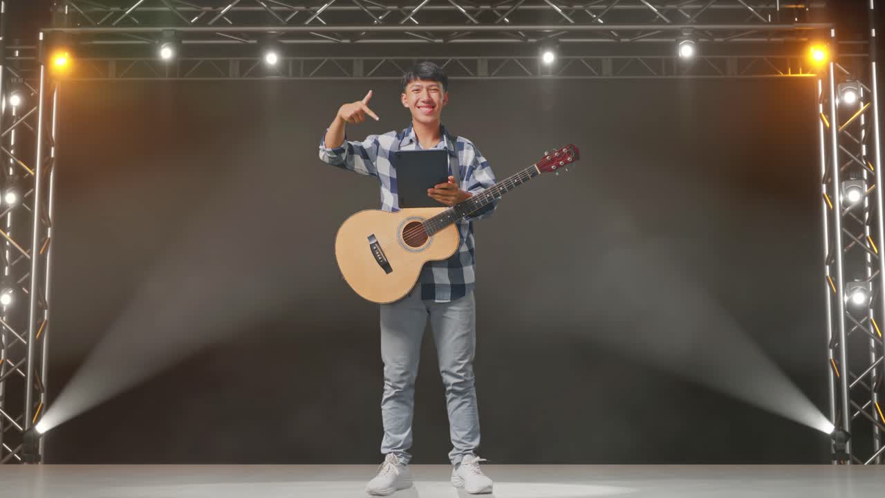 무대에서 서 있는 동안 손에 태블릿을 가리키는 기타를 들고 있는 아시아 십대의 몸이 가득 차 있습니다.