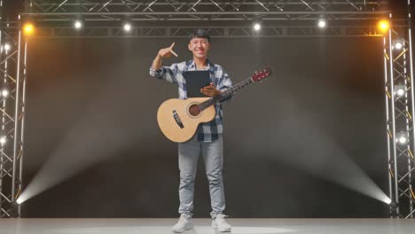 cuerpo lleno de adolescente asiático con guitarra apuntando a una tableta en la mano mientras está de pie en el escenario