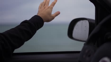 A man&#039;s hand is catching the rain out while driving a car on a rainy day