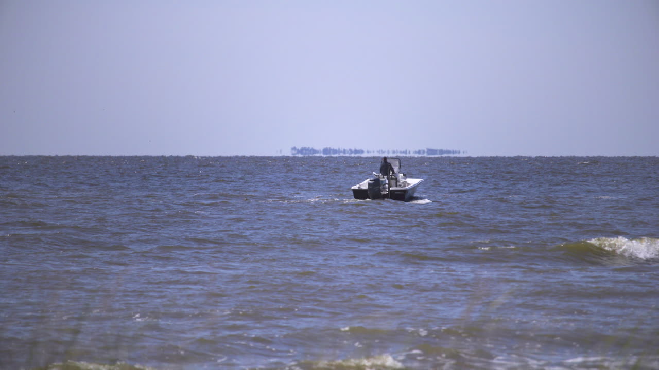 A small bass boat floats in choppy water off of the Gulfport Harbor in the Gulf of Mexico. A male figure is driving a boat in choppy water during the day.