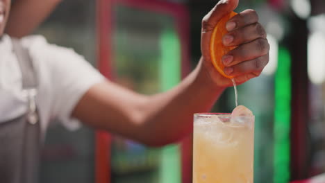 barman squeezes orange half into cocktail in bar closeup. african american barkeeper adds fresh citrus ingredient to drink in nightclub. party beverage