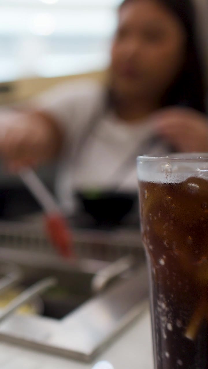 Un bicchiere alto di cola ghiacciata è a fuoco mentre una donna mangia sullo sfondo.