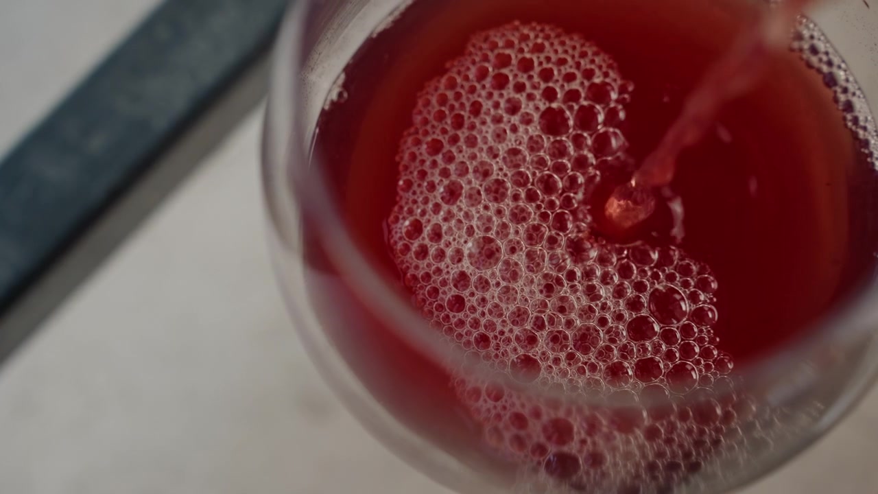 Liquido rosso riempie un bicchiere formando bolle e schiuma mentre viene versato. Lo sfondo include un oggetto scuro sfocato su una superficie chiara. L'illuminazione evidenzia la texture del fluido rosso in una vista ravvicinata.