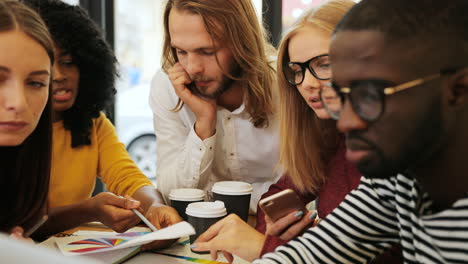 close-up view of multiethnic coworkers viewing graphics and taking notes sitting at a table in a cafe