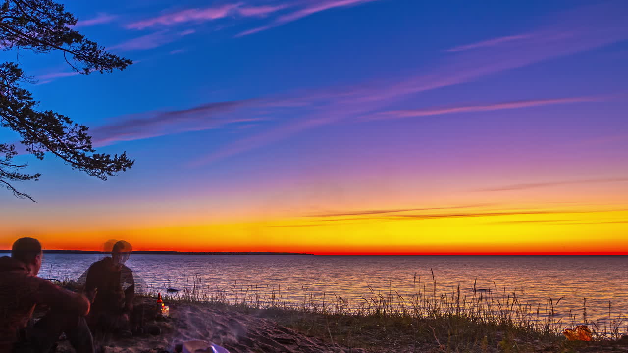 Stunning sunset over a serene lake with people at a camp fire, vibrant colors and tranquil atmosphere, timelapse