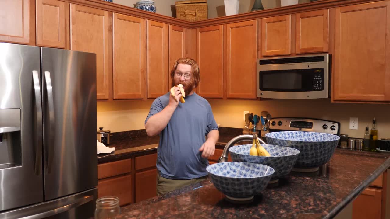 un hombre de pelo rojo comiendo un plátano en una cocina.