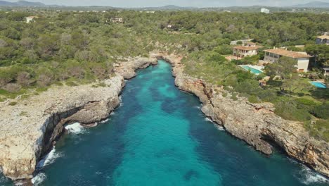 an aerial view of a dramatic rocky inlet with vibrant turquoise water surrounded by cliffs and dense forest. elegant villas with swimming pools are perched along the cliffside.
