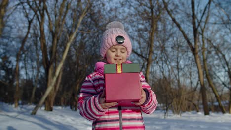 Lächelndes-Kind-Im-Verschneiten-Winterwaldpark,-Das-In-Die-Kamera-Blickt-Und-Eine-Geschenkbox-Mit-Weihnachtsgeschenken-Hält