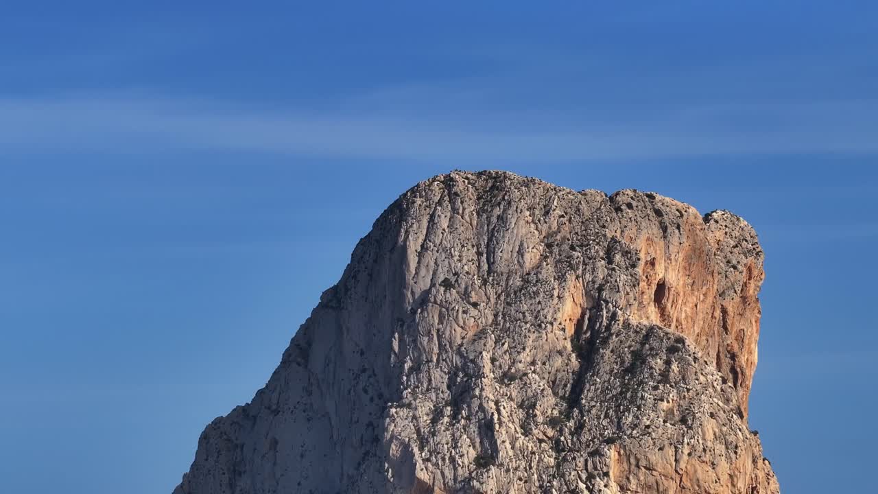 Camera tilt reveals Calp's marina filled with boats, then slowly rises to showcase the imposing rocky cliff of Peñón de Ifach with Mediterranean blue waters and rugged vegetation, real time