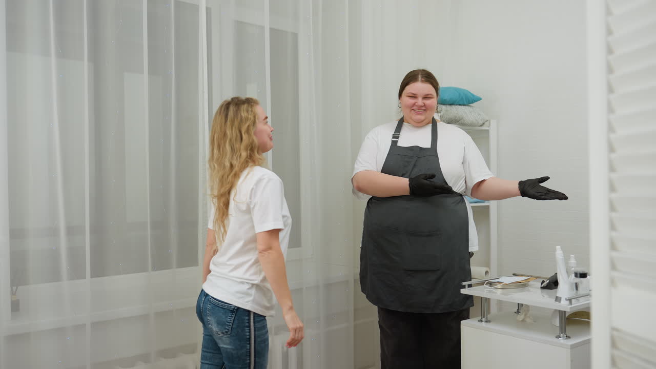 Beautician in apron stands with warm smile while welcoming client into clean modern salon. Bright interior with curtains, cosmetic tools, and organized shelves set cozy professional atmosphere