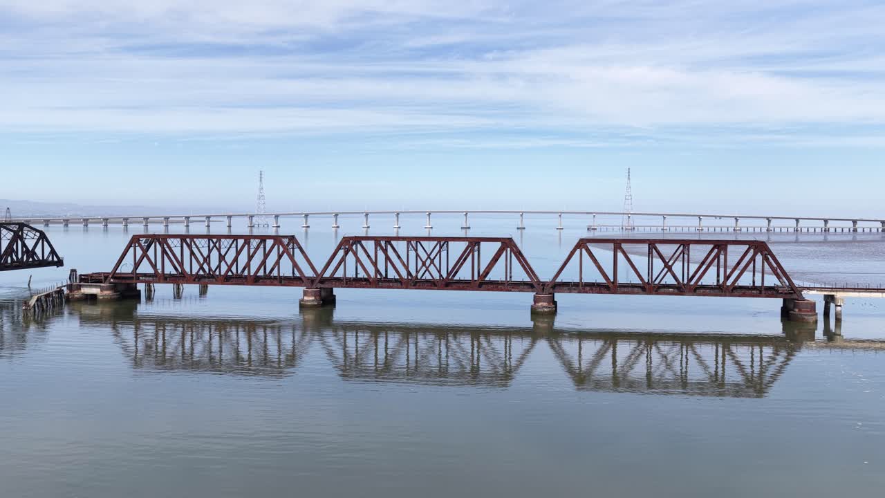 Aerial footage moving sideways along the abandoned Dumbarton Rail Bridge, revealing its rusty truss structure and the open swing section over the calm waters of the San Francisco Bay