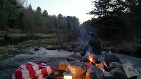 man fishing by a campfire with a lantern and blanket in a forest at twilight, surrounded by trees and the tranquil sounds of nature