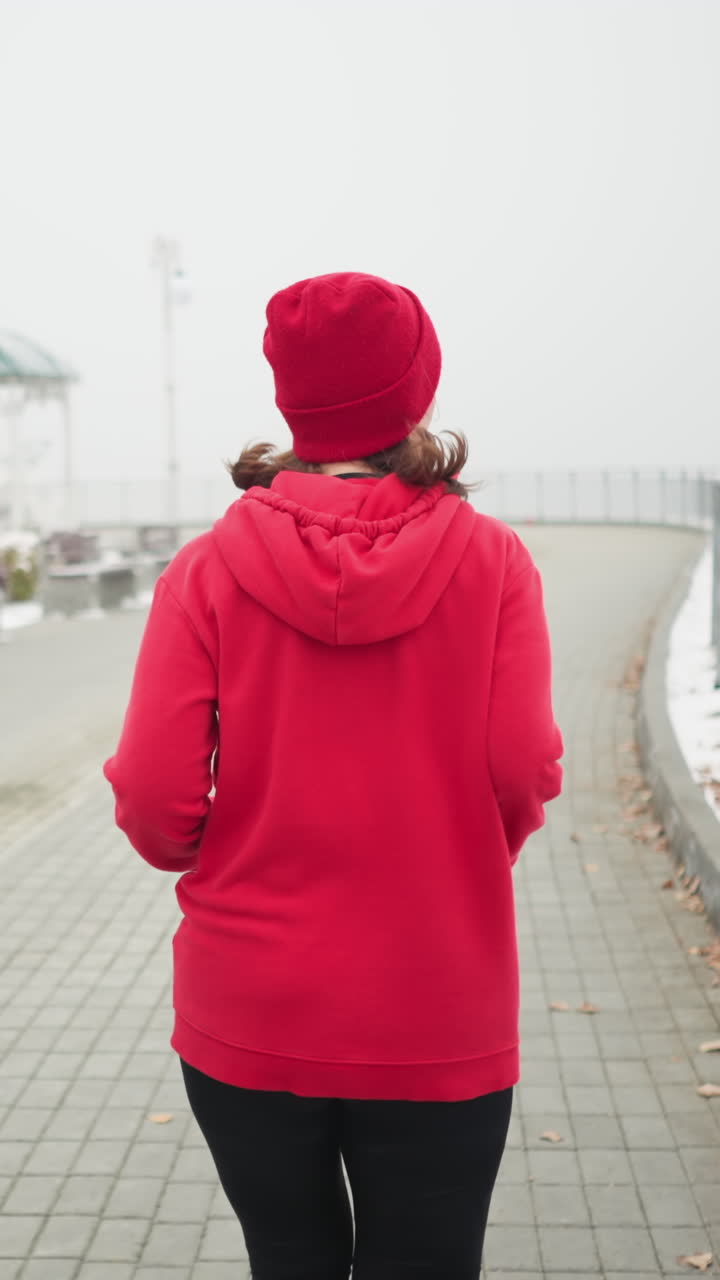 back view of woman jogging in red beanie and jacket along snowy pathway by iron railing and benches in foggy winter park with distant bridge and snow covered trees in cold fitness routine