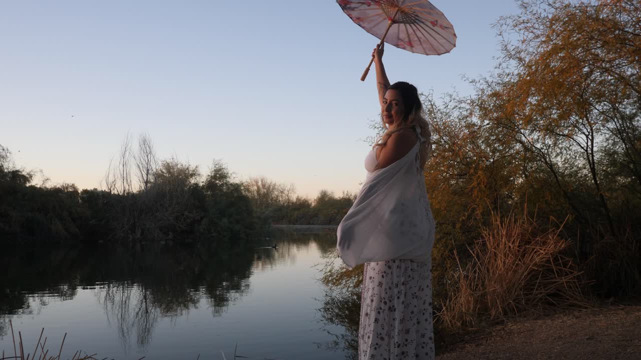 Graceful woman spins umbrella during sunset next to a calm pond.