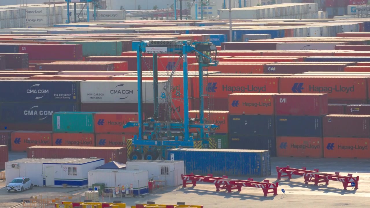 Industrial straddle carrier lifting and transporting a shipping container across a busy commercial dockyard filled with stacked cargo, representing global logistics and international trade