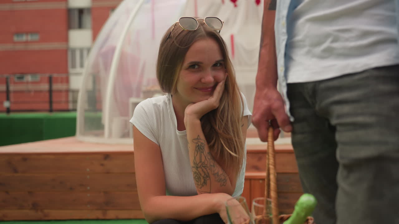 white woman and man on rooftop turf, smiling crouched beside wicker basket and clear dome, casual clothes with sunglasses and tattoo, playful gaze and gentle touch, warm daytime light, urban terrace