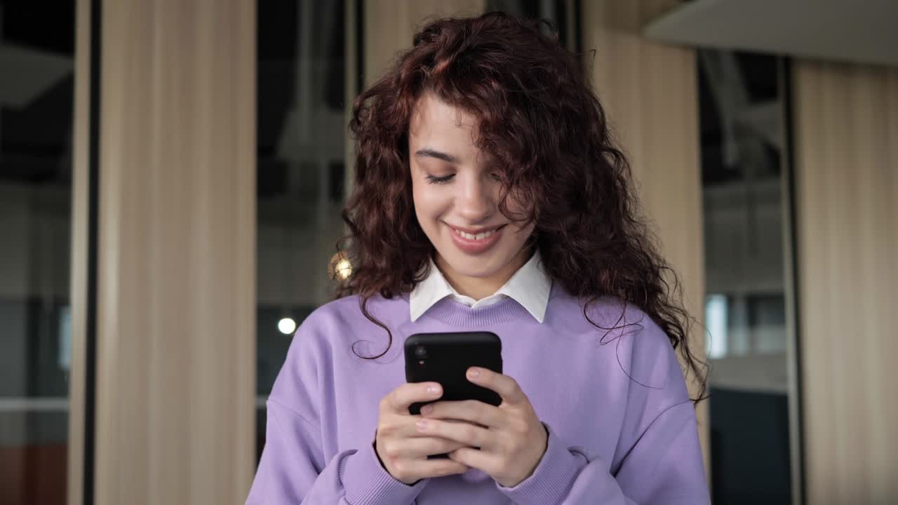 mujer independiente con cabello rizado escribiendo un mensaje en el teléfono móvil en la oficina. joven diseñadora en un suéter púrpura hablando por teléfono. cerca de las manos de la mujer joven usando teléfono inteligente en el interior.