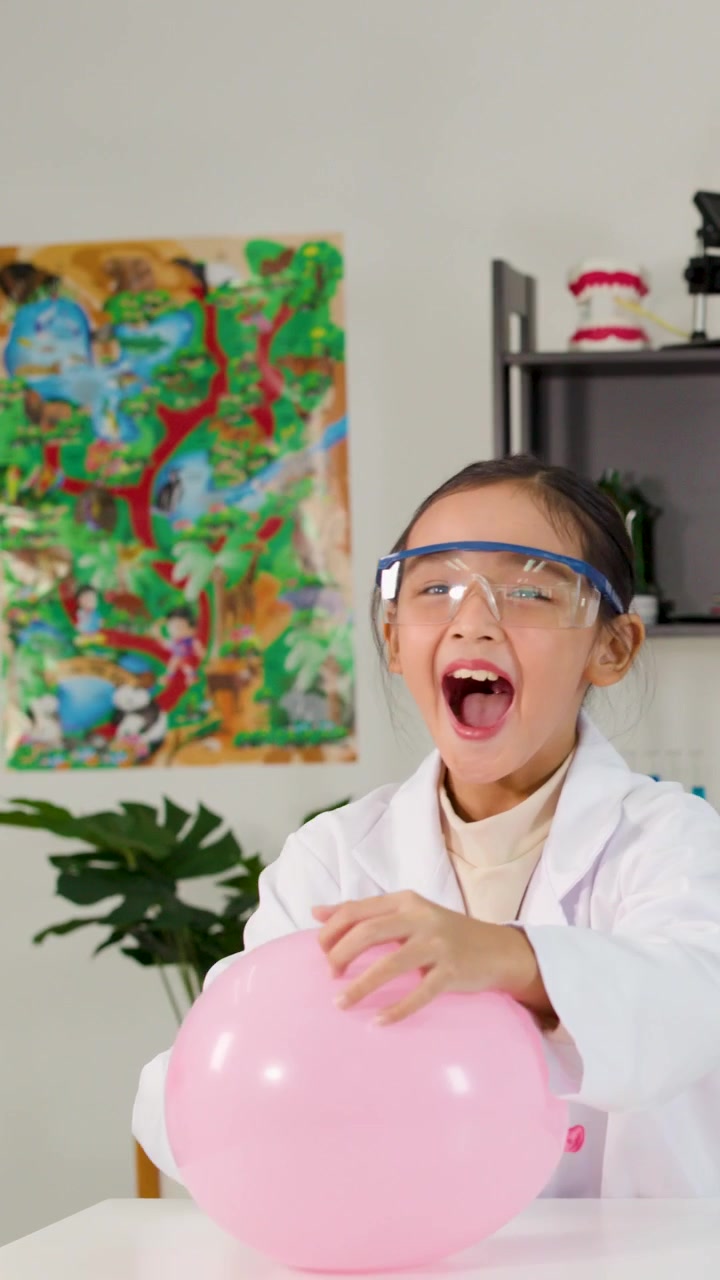 A young girl wearing safety goggles pops a pink balloon and laughs in a classroom