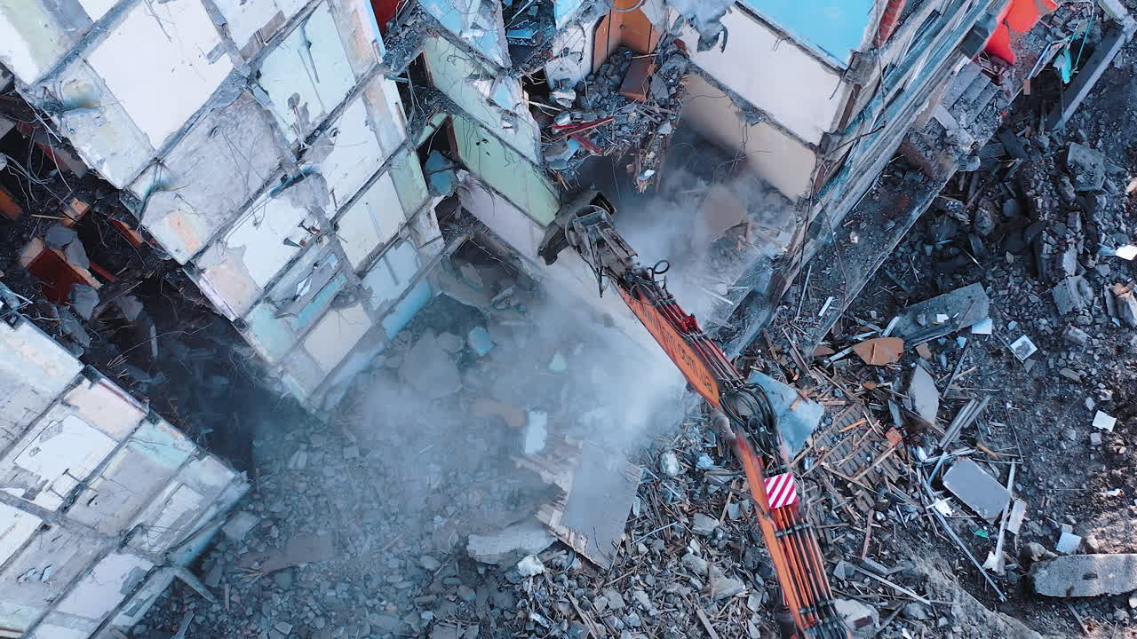 Huge building being demolished with the help of a tractor. Excavator with a long claw breaking walls of the ruined building. View from the above.