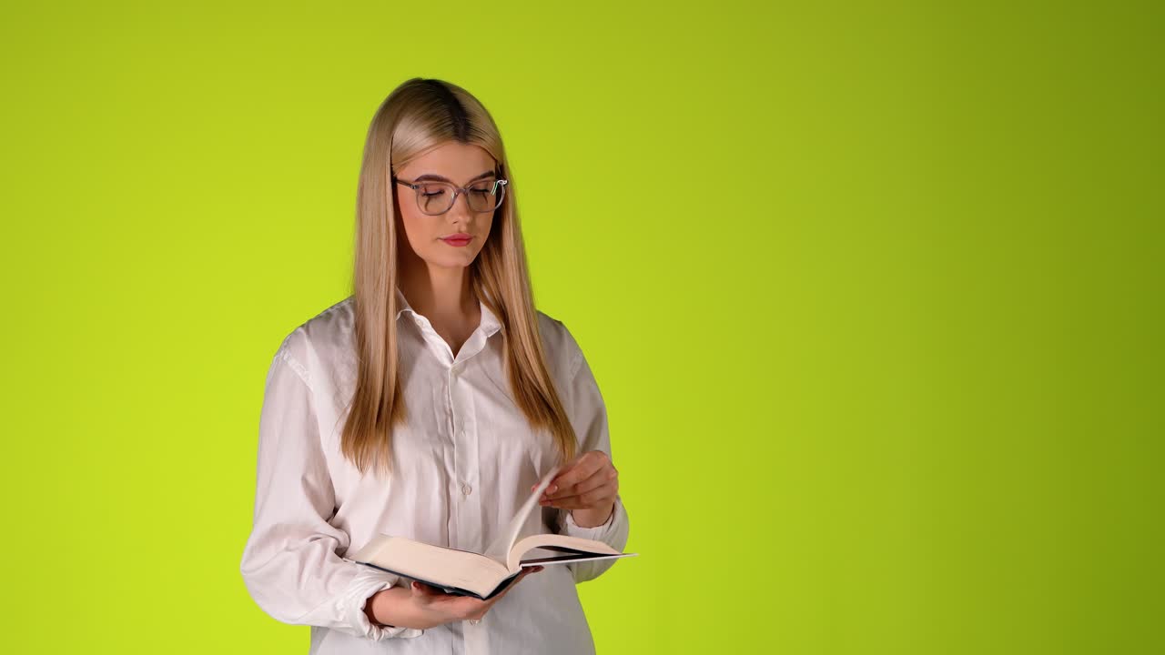 Giovane donna bionda intellettuale che legge un libro in piedi, ritratto in studio colorato