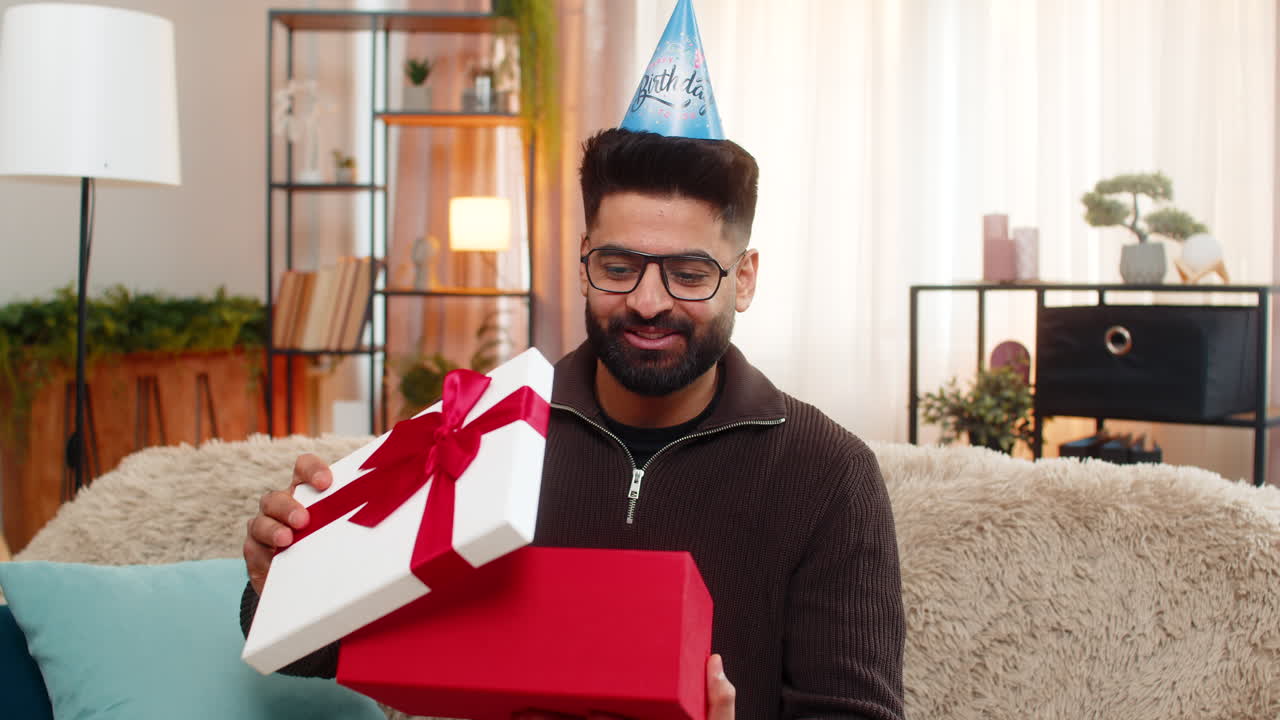 Indian man on sofa opens birthday gift with smile showing joy and surprise in cheerful celebration