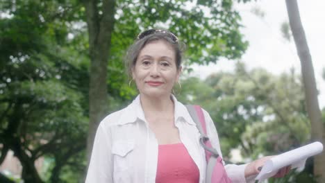 Senior-woman-holding-a-paper-looks-and-smiles-at-the-camera-outdoors-with-trees-in-the-background,-radiating-experience-and-confidence