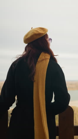 joven con el cabello rojo ondulado en un abrigo negro, caminando hacia una valla de madera, descansando su mano en ella, disfrutando de la vista panorámica de un río y follaje seco