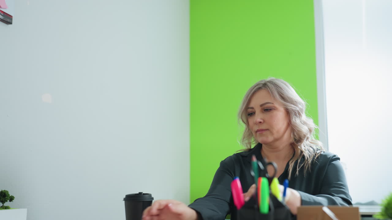 Focused woman opens laptop in bright modern office with green accent wall, preparing to begin remote work session with coffee cup nearby and colorful stationery on organized desk