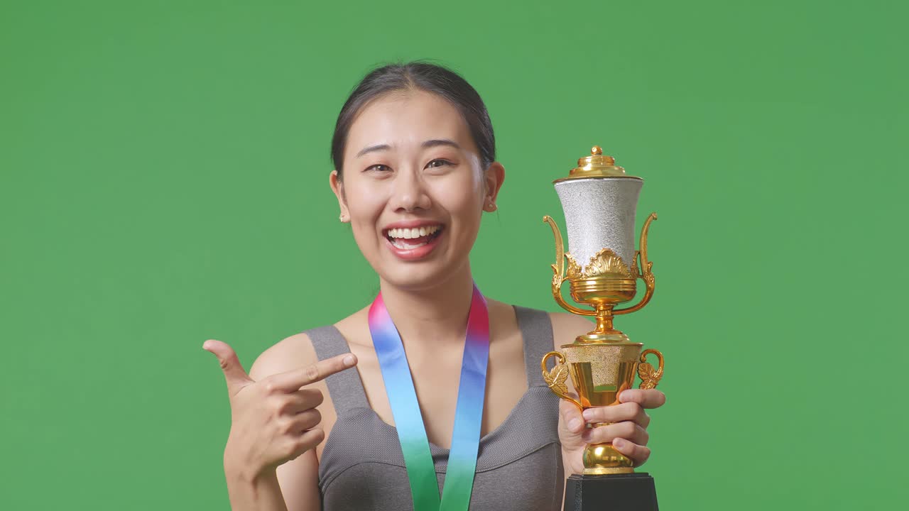 금메달을  아시아 여성이 손에 금 트로피를 가리키며 스튜디오의 녹색 스크린 배경에서 첫 번째 우승자로 자랑스러워합니다.
