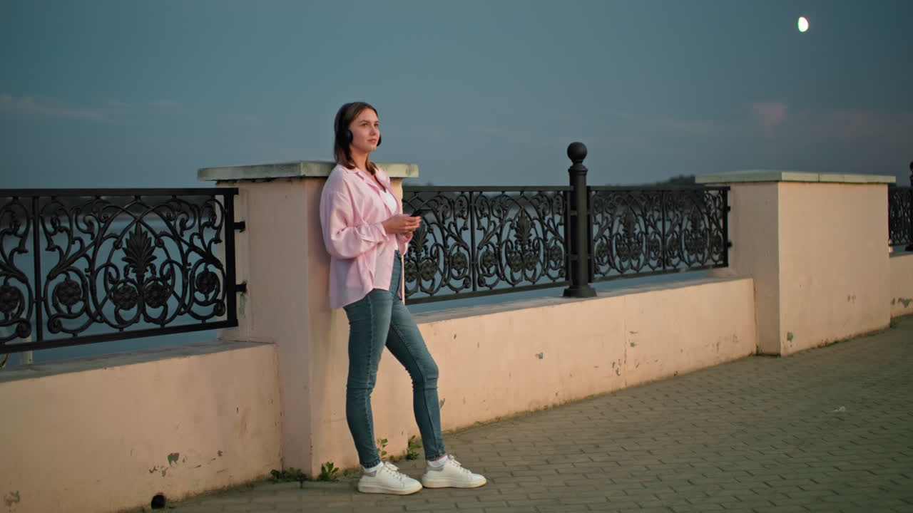 mujer de pie al aire libre al anochecer, apoyándose en una valla de hierro decorativa, usando auriculares y sosteniendo un teléfono, con una suave iluminación nocturna y la luna visible en el fondo