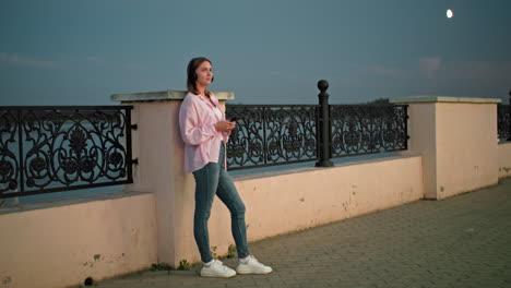 woman standing outdoors at dusk, leaning against a decorative iron fence, wearing headphones and holding a phone, with soft evening lighting and moon visible in the background