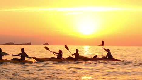 traveling shot of an expedition of kayaks cross in front of the sunset at the goldenhour in slowmotion at playa escondida - ibiza