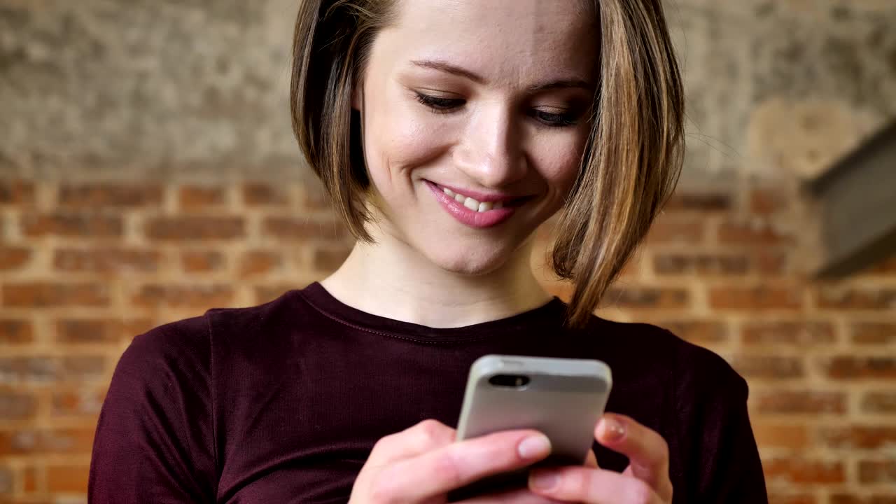 Giovane ragazza bella sta leggendo un messaggio sul suo smartphone, è sorpresa, concetto di comunicazione, sfondo di mattoni.