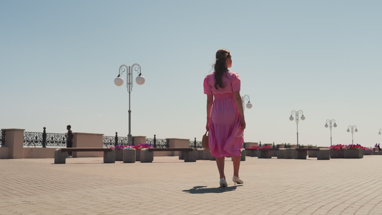 Donna in abito rosa che passeggia sul lungomare con gonna svolazzante e passo sicuro, ciottoli illuminati dal sole, lampioni decorativi, ringhiera in lontananza, ciclisti di passaggio, energia estiva, filmato lifestyle
