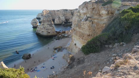 Tiro-Inclinado,-Vista-Panorámica-De-Personas-Bañándose-En-La-Costa-De-La-Playa-Del-Túnel-De-Roca-En-Algarve,-Portugal,-Olas-Golpeando-La-Formación-Rocosa-En-El-Fondo