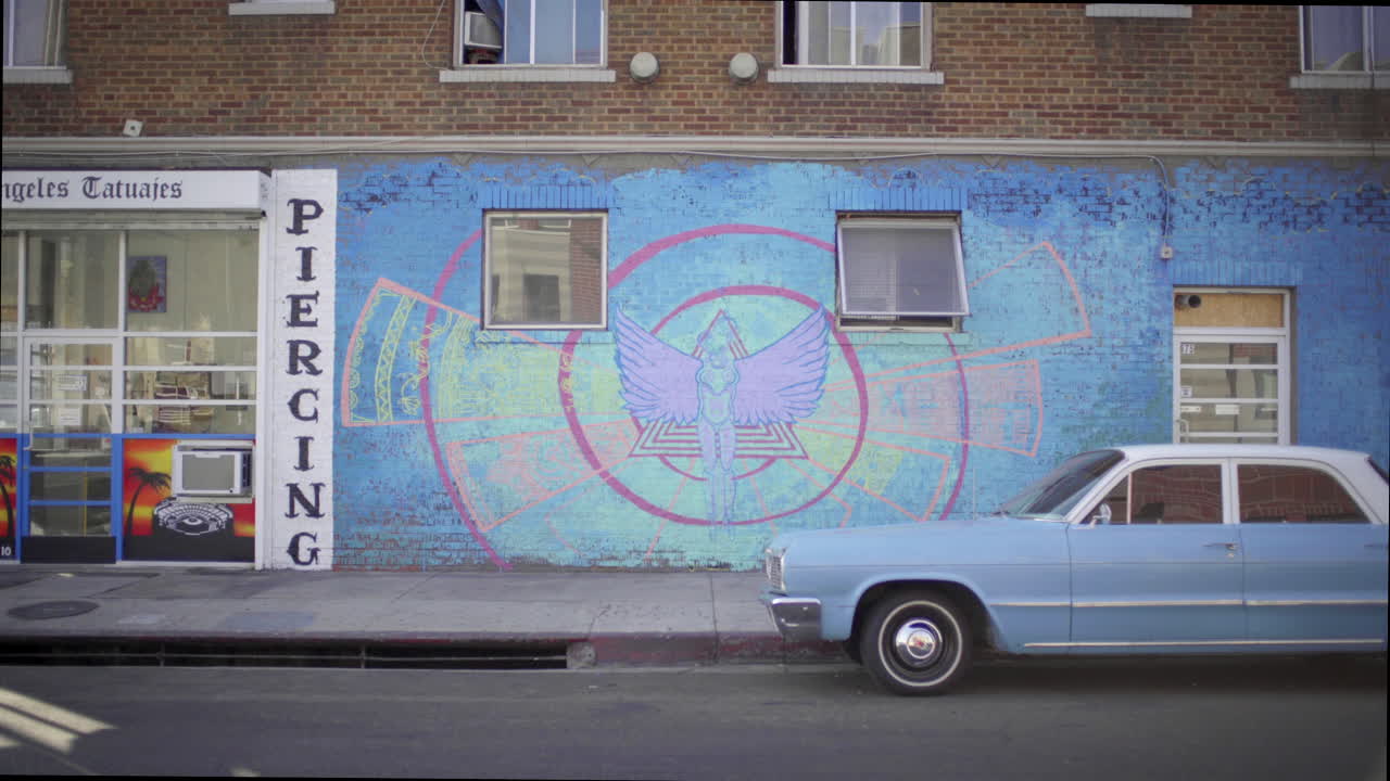 A vibrant blue mural with a winged figure on a brick wall, next to a classic light blue car parked on an urban street