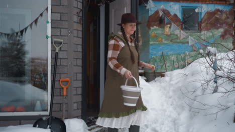 pet lover wearing hat and long gown scoops fresh snow into empty bucket walking out into snowy yard, her dog by side under gray sky in forest clearing, surrounded by frost covered branches