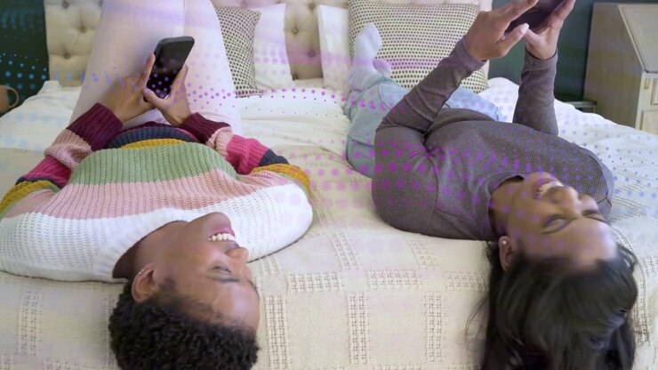 Due donne a letto che interagiscono con i loro telefoni e mostrano gli schermi, condividendo contenuti social con reazioni animate.