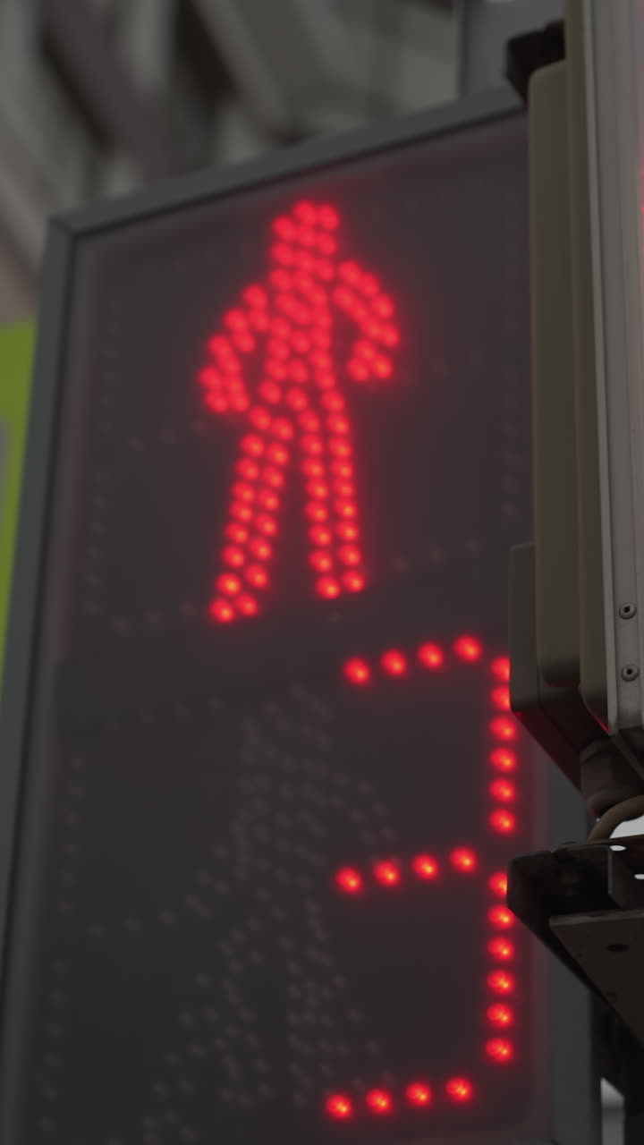 vista en primer plano de un semáforo digital para peatones que muestra una señal roja y un temporizador de cuenta atrás, el semáfero está posicionado al lado de una señal de cruce peatonal, con un edificio de la ciudad