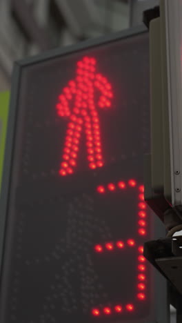 vista en primer plano de un semáforo digital para peatones que muestra una señal roja y un temporizador de cuenta atrás, el semáfero está posicionado al lado de una señal de cruce peatonal, con un edificio de la ciudad