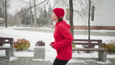 side view of woman jogging along snowy park pathway in red hoodie and black leggings, surrounded by serene winter scenery with benches, bushes and white building in the background