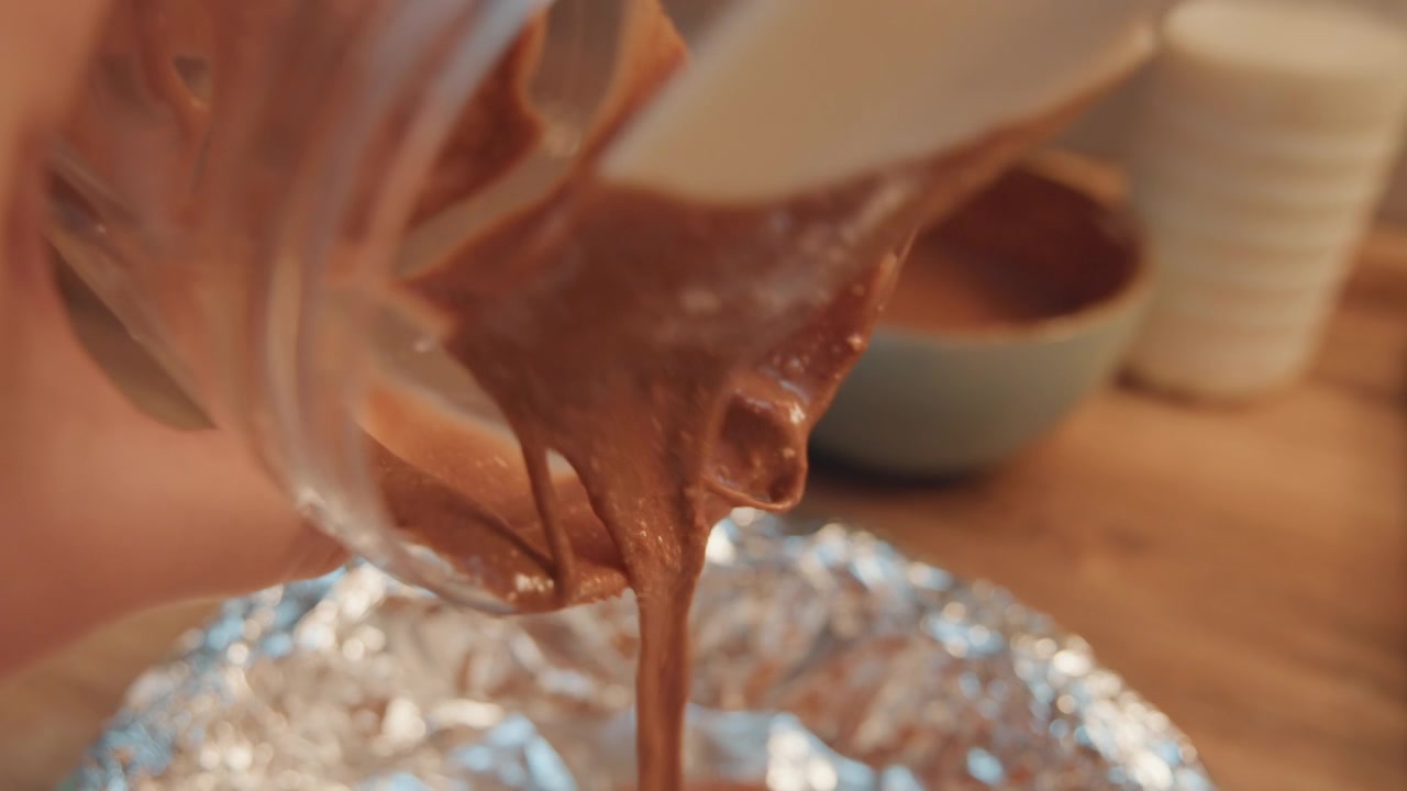 Mani dall'alto versano impasto al cioccolato da un barattolo in una teglia foderata di alluminio, spatola che raschia il vetro mentre ciotole da cucina calde sono sullo sfondo, momento di preparazione intimo che evidenzia tecnica e ritmo.