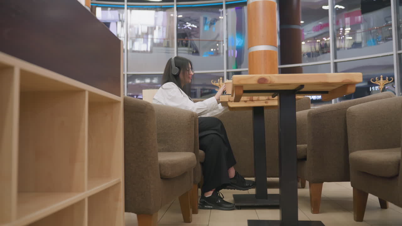side view of focused woman wearing headphones working on tablet in cozy cafe setting with warm lighting and modern mall interior visible through large glass windows in background during evening