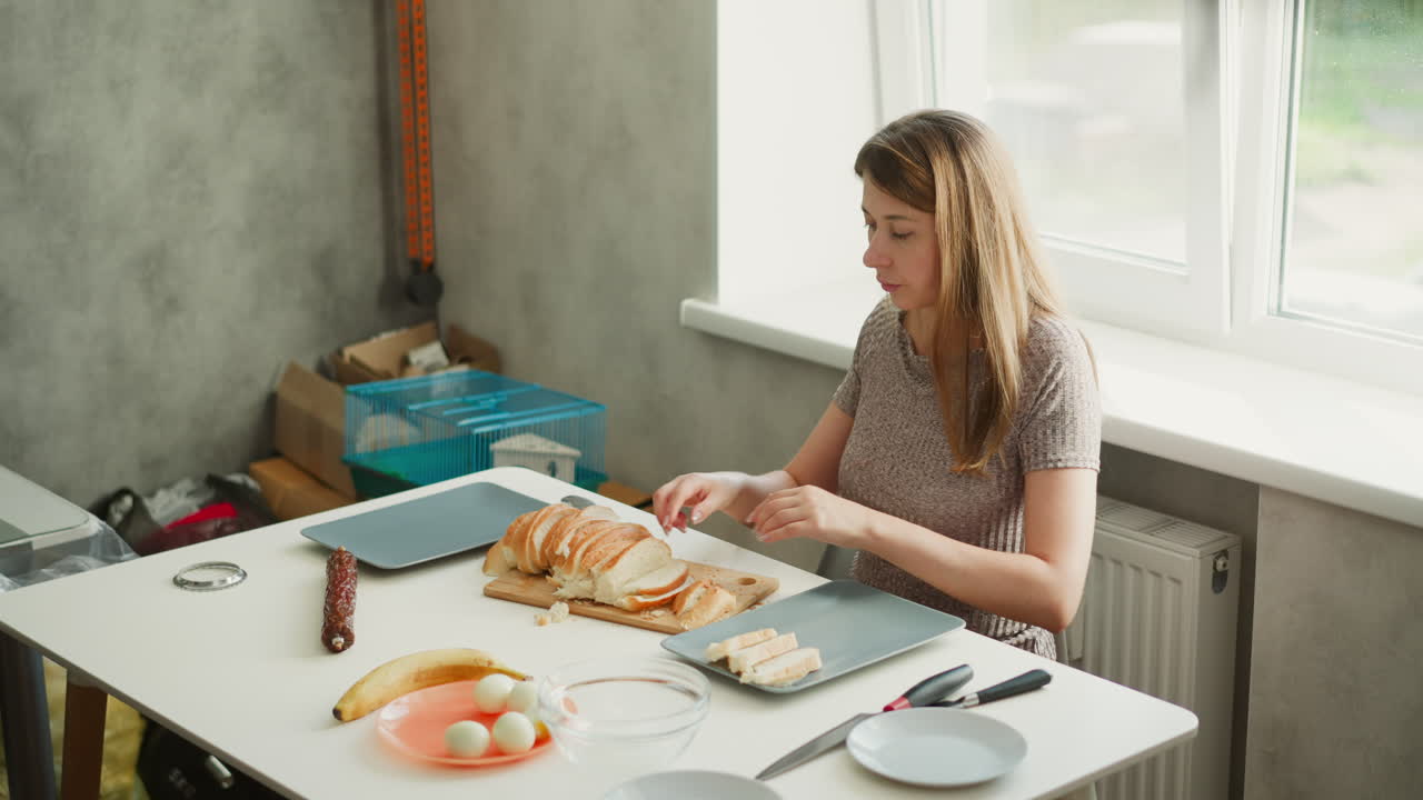 Medium shot of pastry chef placing sliced bread on plate with careful attention to process, table set with eggs, banana, bowls and knife under bright window light creating focused culinary scene