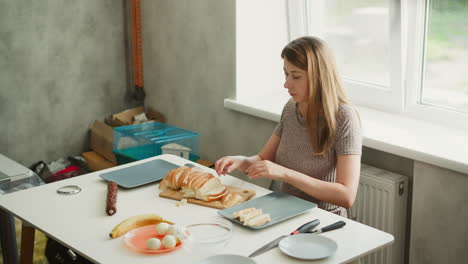 medium shot of pastry chef placing sliced bread on plate with careful attention to process, table set with eggs, banana, bowls and knife under bright window light creating focused culinary scene